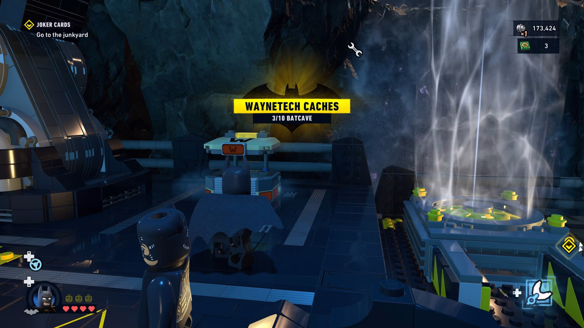 In the game 'LEGO Batman Legacy of the Dark Knight,' the player controls Batman inside the Batcave, with on-screen text reading 'WAYNETECH CACHES 3/10 BATCAVE' and 'JOKER CARDS Go to the junkyard.'