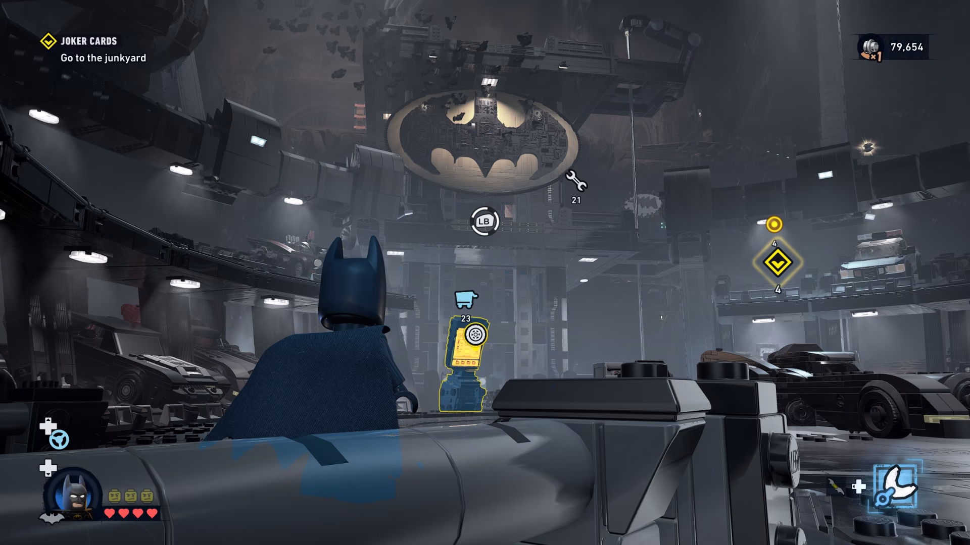 In 'LEGO Batman: Legacy of the Dark Knight' video game, Batman stands in the Batcave surrounded by vehicles with a mission objective reading 'Go to the junkyard.'